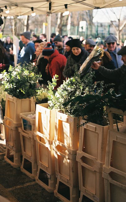 A busy outdoor flower market scene featuring wooden display crates filled with lush, green floral arrangements, including various types of foliage and flowering plants. The bouquets are wrapped in cle