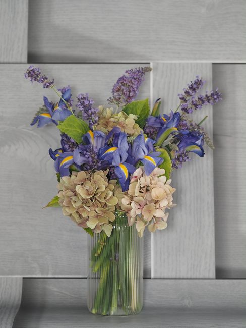 A tall, clear glass vase filled with an arrangement of fresh flowers, including purple irises with yellow accents, soft pink hydrangeas, and delicate lavender sprigs, set against a light gray wooden b