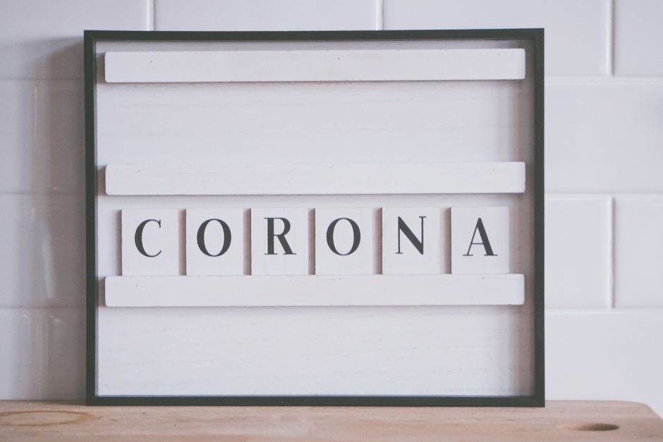 A black-framed white letter board with five horizontal white wooden slats, placed on a wooden surface against a glossy white tiled wall. The board displays the word 'CORONA' spelled out with individua