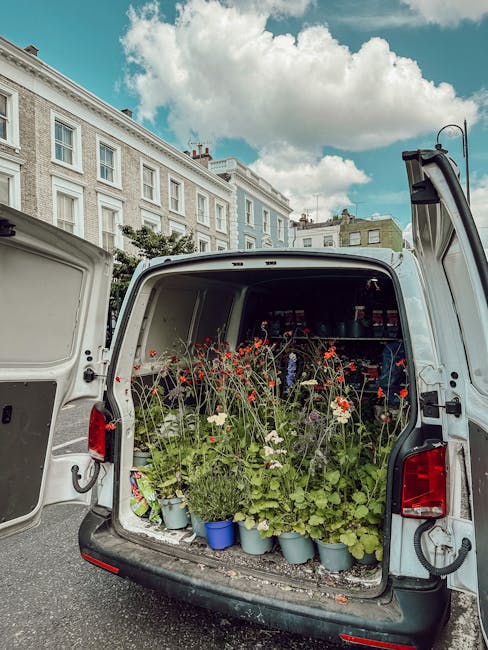 The open rear of a white delivery van filled with potted flowers and plants, including red poppies, white daisies, and assorted green foliage, arranged in blue and gray pots. The van is parked on a st