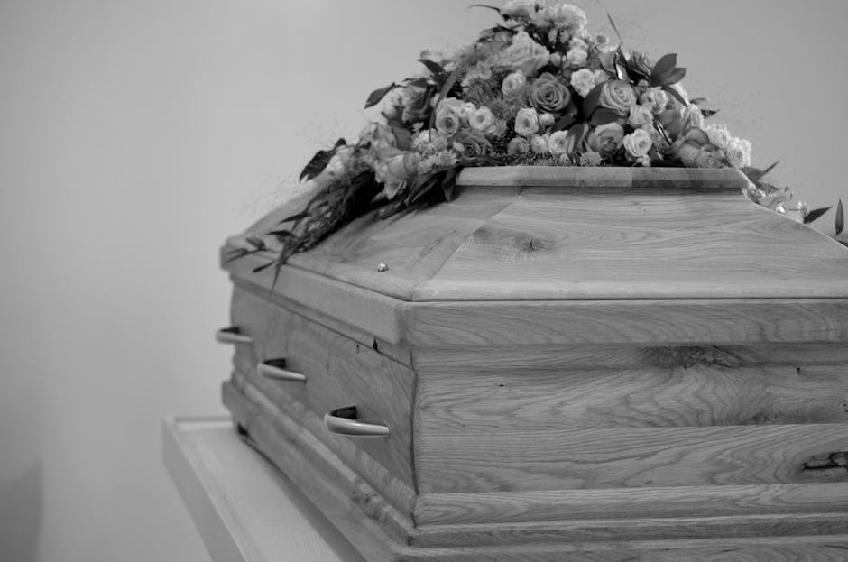 A wooden casket with a smooth, polished surface and natural wood grain visible, draped with a large, elegant floral arrangement consisting of roses, carnations, and greenery in soft pastel shades. The