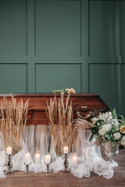 A floral arrangement featuring white roses, peonies, and various greenery placed in a white vase on a table, with a backdrop displayed in a soft green wall. Surrounding the vase are tall pampas grass