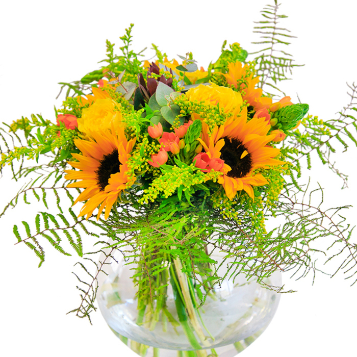 The spirit of Shadwell pulses through this exuberant bouquet, where flower artistry and local colour unite in a vivid display. Towering sunflowers form the heart of the arrangement, their expansive golden petals and textured brown centres reminiscent of sunlight glancing off the Thames at waterside parks. They're joined by elegant golden yellow garden roses, their tightly wrapped petals unfurling in sophisticated whorls, giving depth and an ageless, romantic touch. Crimson Hypericum berries sparkle amid the blossoms, vibrant against the lush tapestry of mixed foliage, like treasured finds in vibrant Watney Market stalls. Feathery fern fronds cascade gently, blending with sprigs of yellow goldenrod and wisps of light green foliage that whisper of Shadwell's landscaped gardens. Cool, blue-tinted eucalyptus leaves peek from between the flowers, infusing modern texture and understated fragrance. The bouquet is carefully poised within a clear, spherical glass vase, its water gleaming and stems elegantly displayed, creating a clean, contemporary finish. Whether serving as a radiant centrepiece for a riverside dinner, an uplifting gift for someone dear, or a cheerful accent in a Shadwell home, this arrangement captures the season's natural beauty and the local florist's dedication to charm and craftsmanship.