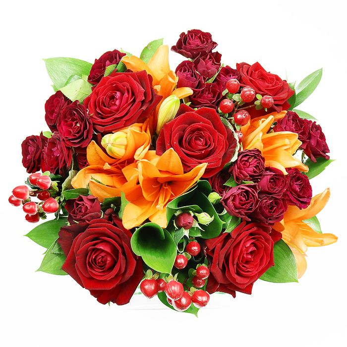 From an overhead perspective on a white backdrop, this opulent bouquet reads as a perfectly rounded study in autumnal romance. Prominent are several large, fully opened deep red roses whose velvety, layered petals create soft shadows and delicate highlights; these classic blooms embody passion and a timeless elegance. Interspersed among them are brilliant orange Asiatic lilies, their trumpet-shaped petals and visible stamens adding structural drama-some are fully unfurled, revealing speckled inner throats, while others remain poised as elongated buds, suggesting movement and unfolding life. Threaded through the arrangement are clusters of deep burgundy spray roses, their miniature rosettes offering a refined, plush texture and intensifying the bouquet's jewel-like depth. Bright red hypericum berries appear in small clusters, their glossy, spherical forms providing a cheerful, tactile contrast to the soft petals. Broad, glossy green leaves-reminiscent of salal or ruscus-peek from beneath the blooms, framing the composition and cooling the rich reds and oranges with fresh, natural green. The soft, even lighting highlights petal veins and berry gloss without casting heavy shadows, giving the bouquet a polished photographic clarity while preserving the sense of handcrafted artistry. As a floral piece suited to anniversaries, congratulations, or a heartfelt thank-you, it would sit beautifully on a Shadwell coffee table or be delivered for celebration near Cable Street and Wapping; the arrangement balances warmth, texture, and scent in a manner that feels both generous and carefully composed.